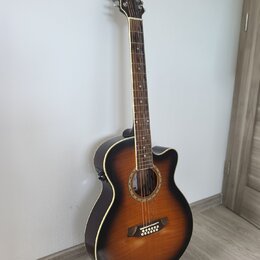 Akustická kytara s cutawayem v tmavě hnědém sunburst provedení. Na hlavě je viditelné logo "Cort". Nástroj je umístěn na černém stojanu u bílé stěny.