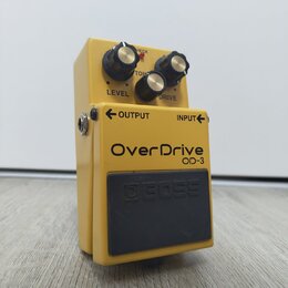 Žlutý kytarový efekt s černým spínacím pedálem. Na horní části jsou tři černé ovládací knoflíky s nápisy LEVEL, TONE a DRIVE. Na těle je uveden název…