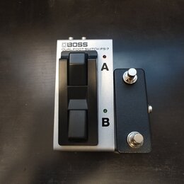 Stříbrný dvoupedálový spínací pedal s nápisem BOSS DUAL FOOT SWITCH FS-7. Vedle něj je umístěn malý černý box se dvěma kovovými přepínači. Na stříbrn…