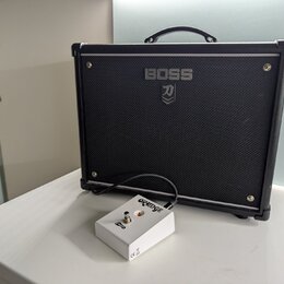 Černý kytarový kombo zesilovač s logem BOSS a symbolem katany pod ním. Před zesilovačem je umístěn malý bílý efektový pedál s nápisem ORANGE a označe…