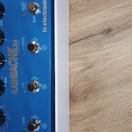 Modrý kytarový efekt s bílým rámem. Na horní straně je nápis FLASHBACK DELAY AND LOOPER a logo tc electronic. Zařízení má čtyři černé otočné knoflíky…