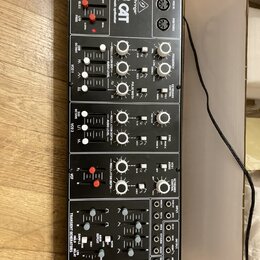 Černý elektronický syntetizátor s nápisem "behringer CAT" a přívstkem "electronic music synthesizer". Přední panel obsahuje množství bílých posuvníků…