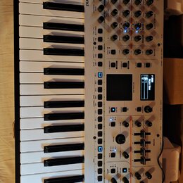 Bílý syntetizátor s klaviatura značky Roland, model GAIA 2. Přístroj má numerous ovládací prvky v podobě posuvníků, otočných knoflíků a tlačítek, vče…