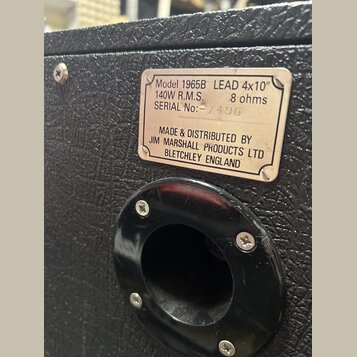 Detail zadní strany kytarového reproboxu s černým texturovaným povrchem. Na kovovém štítku je uveden nápis: Model 1965B LEAD 4x10", 140W R.M.S., 8 oh…