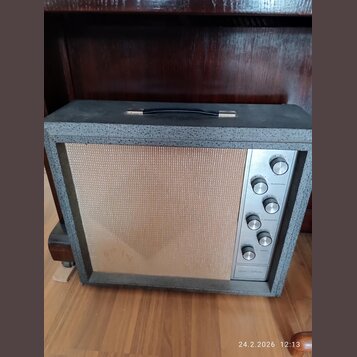 Kytarový zesilovač v šedém texturovaném potahu s béžovou mřížkou. Na pravé straně je kovový ovládací panel s knoflíky a nápisy MICROPHONE, INSTRUMENT…