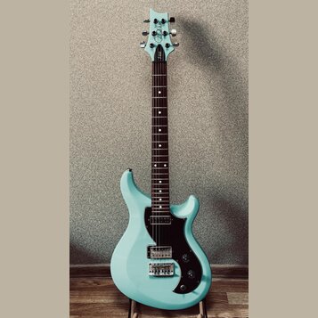 Světle modrá elektrická kytara s černým pickguardem a hnědým hmatníkem. Na hlavě je bílé logo "Cort" a nápis "Veta". Kytara má dva humbuckery, jeden…