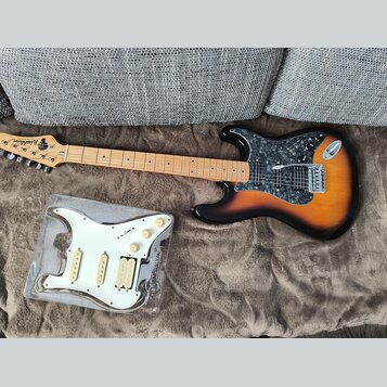 Elektrická kytara s tělem v provedení sunburst a hnědým hřebenatým pickguardem. Na hlavě je viditelné logo Washburn. Vedle kytary leží v plastovém ob…