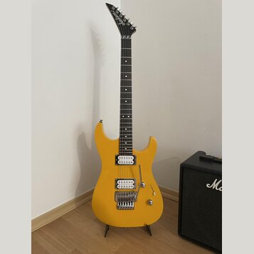 Žlutá elektrická kytara s tmavým hnedým hmatníkem a bílými tečkovanými značkami. Na hlavě je viditelné logo Jackson. Kytara má dva humbuckery, tremol…