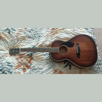 Akustická kytara s hnědým povrchem v provedení sunburst a černým plektrem. Na hlavě hрифu je bílý nápis Fender. Nástroj vykazuje známky běžného použí…