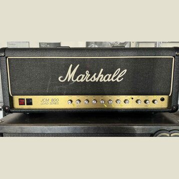 Kytarový zesilovač značky Marshall, model JCM 800 LEAD SERIES. Má černý kryt s bílým logem Marshall a zlatý ovládací panel s řadou otočných knoflíků.…