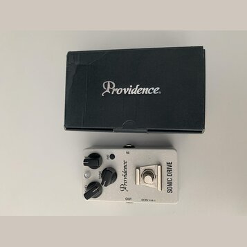 Kytarový efekt pedál stříbrné barvy s nápisy Providence a SONIC DRIVE. Pedál má tři černé ovládací knoflíky s popisky FAT, TONE a LEVEL. Vedle je umí…