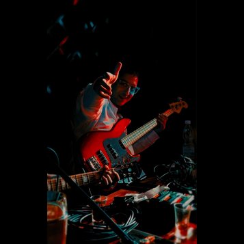 Fotografie zachycuje muže s červenou elektrickou basovou kytarou. Nástroj má červené tělo, bílý pickguard a světle hnědý hmatník. Na snímku nejsou vi…