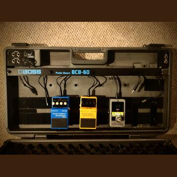 Šedý přenosný kufřík s nápisem BOSS Pedal Board BCB-60, v němž jsou umístěny tři efektové pedály. Zleva je modrý pedál s nápisy Compression Sustainer…