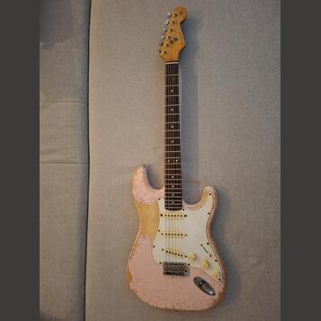 Růžová elektrická kytara typu Stratocaster s bílým pickguardem a hnědým hmatníkem. Lak na těle vykazuje výrazné opotřebení a odrolení v okrajích, kde…