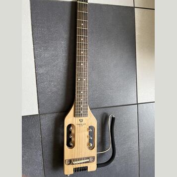 Světle hnědá elektrická kytara s krátkým tělem a dlouhým hnedým hmatníkem. Na těle je černý nápis TRAVELER GUITAR a nad ním logo ve tvaru trojúhelník…