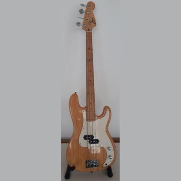 Elektrická basová kytara s přírodním dřevěným povrchem těla a hmatníkem. Na hlavě je viditelné logo "Fender". Instrument má bílý pickguard a čtyři st…