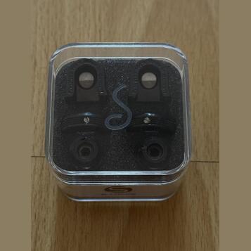 Průhledná plastová krabička s černou pěnovou vložkou obsahuje dva černé kytarové strap locky. Na horních částech jsou vyryty nápisy "earlier" a "guar…