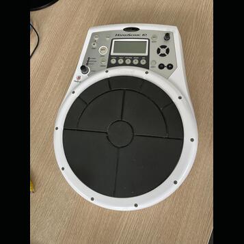 Bílý elektronický perkusní pad Roland HandSonic 10 s velkým černým gumovým playing polem. V horní části je ovládací panel s displejem, několika tlačí…