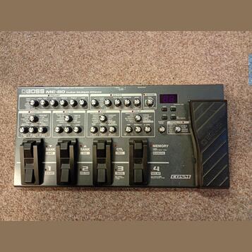 Šedý multifektoped Boss ME-80 Guitar Multiple Effects. Zařízení obsahuje řadu ovládacích knofíků, přepínačů a čtyřistupňové displeje s nápisy jako PR…