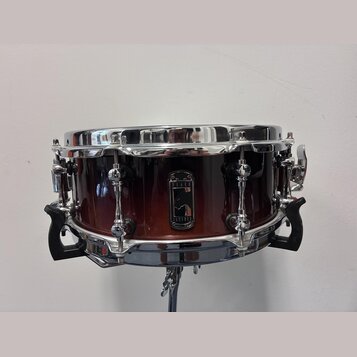 Kytarový snare drum s korpusem v červenohnědém přechodovém odstínu a chromovanými obručemi. Uprostřed korpusu je černý štít s bílým logem a nápisy BL…