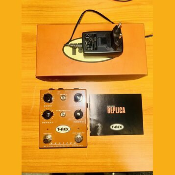 Kytarový efekt pedál v oranžové barvě s nápisy REPLICA, T-REX a BY T-REX ENGINEERING - DENMARK. Zařízení má čtyři ovládací knoflíky, dva přepínače a…