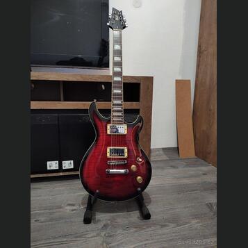Elektrická kytara s červeným gradientem a černým okrajem, umístěná na černém stojanu. Na hlavě hmatníku je bílý nápis Cort. Nástroj má dva humbuckery…