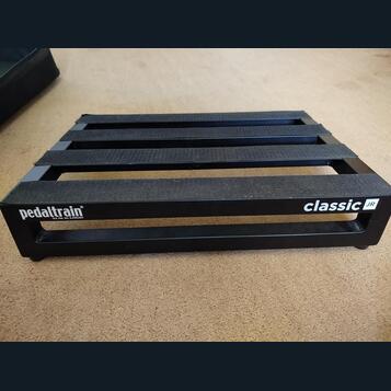 Černý kovový pedalboard s mřížkovou konstrukcí a vložkami z černého textilního popruhu. Na přední hraně je bílý nápis pedaltrain® a pod ním menší tex…
