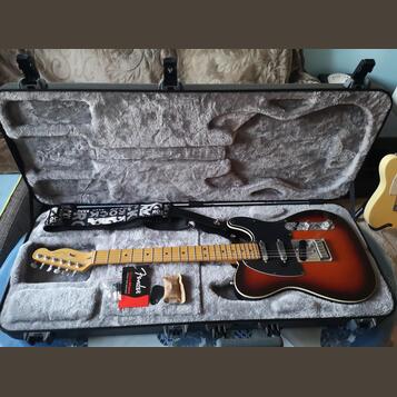 Elektrická kytara s trupem v barevnosti sunburst, černým pickguardem a světle hnědým hnedým hmatníkem. Na hlavě kytary je nápis Fender. Nástroj leží…