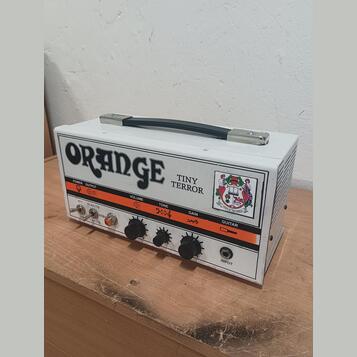 Bílý kytarový zesilovač s nápisem ORANGE a označením modelu TINY TERROR. Na předním panelu jsou čtyři černé ovládací knoflíky s popisky VOLUME, TONE…