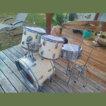 Bílá bicí souprava s chromovaným kováním, sestávající z basového bubnu, dvou tomů a malého bubnu. Na činelech je vidět nápis MEINL a LIVE SOUND. Nást…