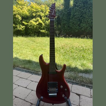 Elektrická kytara s červeným lesklým povrchem korpusu a hnědým hnedým hmatníkem. Na hlavě je bílý nápis Ibanez. Nástroj má dva humbuckery, kobylku s…