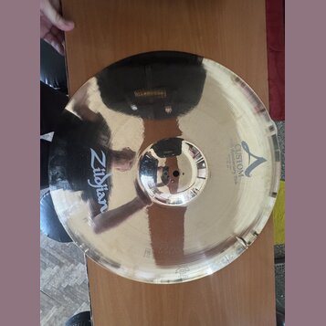 Zlatavý kovový činel s vyznačeným okrajem. Na povrchu je černý nápis Zildjian a logo A s textem CUSTOM 20th Anniversary Ride MEDIUM Thin 22". Povrch…