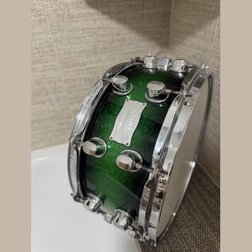 Zelený buben s glitrovým povrchem a chromovaným kováním. Na korpusu je kovový štítek s nápisy „Sultana“, „MAPEX“ a „Custom Series“. Na horním obruči…