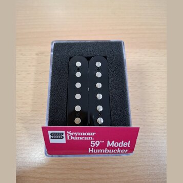 Dvě černé humbuckery s viditelnými bílými póly, umístěné v průhledném plastovém boxu s černou pěnovou vložkou. Na růžovém papírovém pásku je uveden n…