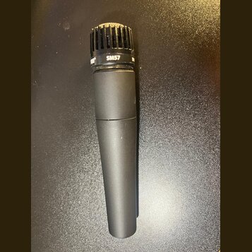 Černý dynamický mikrofon s mřížkou v horní části. Na kroužku pod mřížkou je bílý nápis SM57 a část loga SHURE. Povrch těla vykazuje mírné opotřebení…