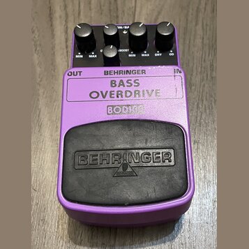 Fialový efekt pedál s černým gumovým spínacím tlačítkem. Na horní části je nápis BEHRINGER BASS OVERDRIVE a model BOD100. Na gumovém tlačítku je vyra…