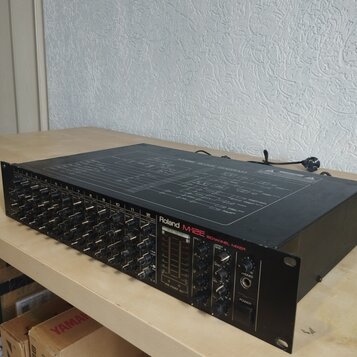 Černý 12kanálový mixážní pult s nápisy Roland M-12E a 12CHANNEL MIXER. Horní kryt obsahuje schéma s názvem M-12E BLOCK DIAGRAM. Zařízení vykazuje mír…