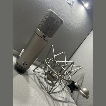 Stříbrný kondenzátorový mikrofon s mřížkou v horní části a kovovým šokabsorberem. Na těle mikrofonu je logo značky a nápis NEUMANN MICROPHONE. V horn…