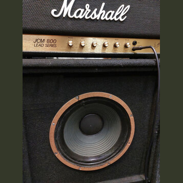 Detail kytarového komba značky Marshall. Horní část tvoří zlatý ovládací panel s nápisem JCM 800 LEAD SERIES a řadou otočných knoflíků. Spodní část u…