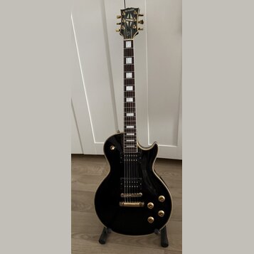 Černá elektrická kytara typu Les Paul se zlatými hardwaremi a bílými blokovými vložkami na hmatníku. Na hlavě je nápis Orville a pod ním menší text b…