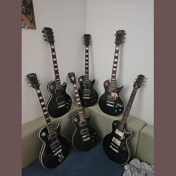 Šest černých elektrických kytar typu Les Paul s bílými bindingy. Na hlavách některých nástrojů je viditelný nápis Epiphone. Kytary mají různé konfigu…