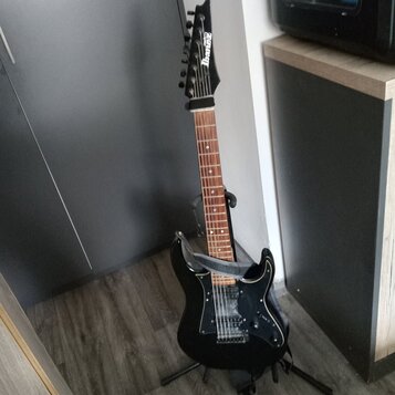 Černá elektrická kytara s hnědým hmatníkem umístěná na stojanu. Na hlavě je bílý nápis Ibanez. Kytara má tři snímače, černý pickguard a připevněný še…