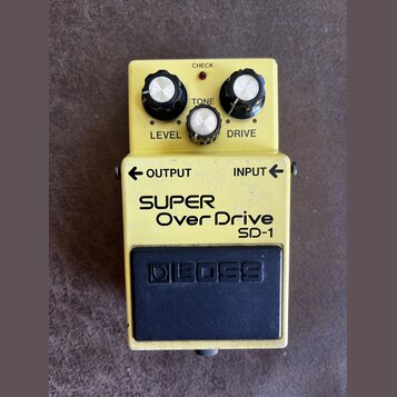 Žlutý kytarový efekt pedál značky BOSS, model SUPER Over Drive SD-1. Na horní části jsou tři černé ovládací knoflíky s bílými vršky a nápisy LEVEL, T…