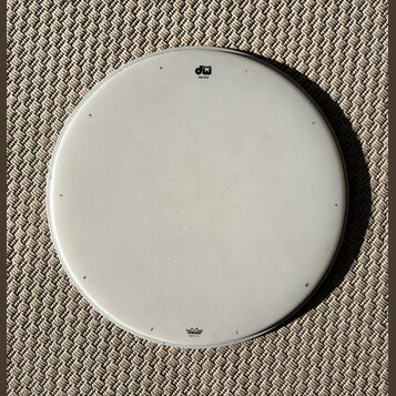 Bílá kruhová bubenová koží v kovovém obruči. V horní části je černé logo „dw drums“ a nápis „coat batter“. Dole se nachází modré logo „Remo“ a text „…