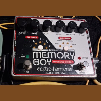 Efekt pedál analogového delay s bílo-černým povrchem a červeným rámem. Na zařízení jsou nápisy DELUXE MEMORY BOY analog delay, electro-harmonix a MAD…