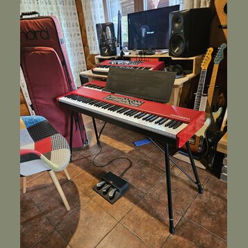 Červený digitální klavír s nápisem nord piano umístěný na černém X-stojanu. Nástroj má připevněný černý notový stojan a k němu patří černý pedál s lo…