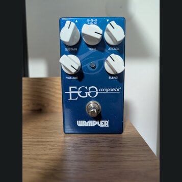 Modrý efekt pedál s nápisy EGO compressor a WAMPLER BUILT IN USA. Zařízení má pět bílých otočných ovladačů s popisky SUSTAIN, TONE, ATTACK, VOLUME a…