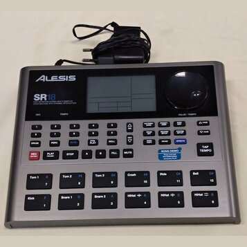 Stříbrný bicí automat značky Alesis, model SR18. Na předním panelu je nápis HIGH DEFINITION STEREO MULTI-SAMPLED DRUM MACHINE WITH DYNAMIC ARTICULATI…