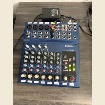 Modrý mixovací pult s nápisem YAMAHA MIXING CONSOLE MG10/2. Horní panel obsahuje různé vstupy a výstupy, včetně konektorů XLR a jack, s popisky jako…