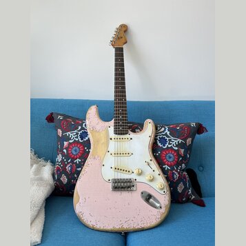 Růžová elektrická kytara typu Stratocaster s bílým pickguardem a hnědým hmatníkem. Povrch instrumentu vykazuje výrazné opotřebení, zejména v podobě o…